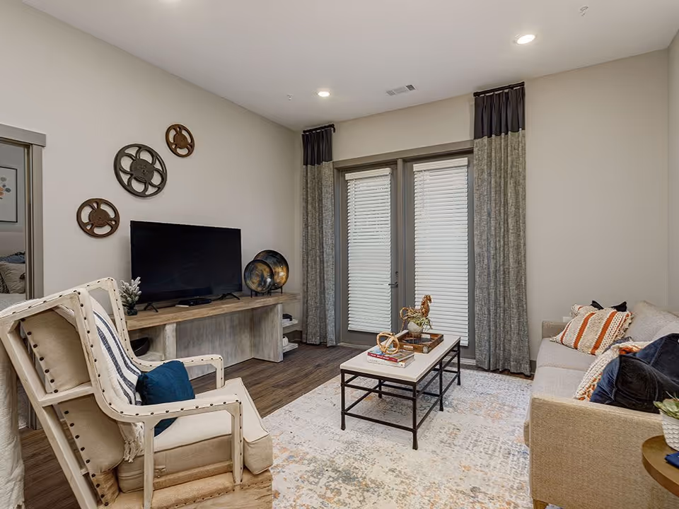 A cozy living room with a beige armchair and a matching sofa adorned with decorative pillows. A rectangular coffee table with books and decorative items sits on a patterned rug. A wooden console holds a flat-screen TV and decorative plates. French doors with closed blinds and long curtains are in the background.
