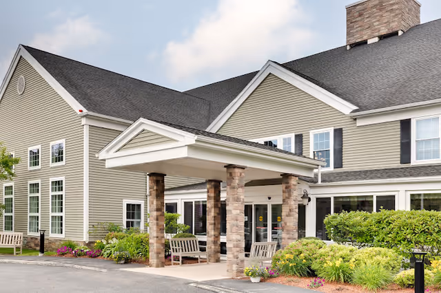 Exterior view of a senior living facility building with beige siding, multiple windows, a covered entrance supported by stone pillars, benches, and landscaped greenery under a partly cloudy sky.