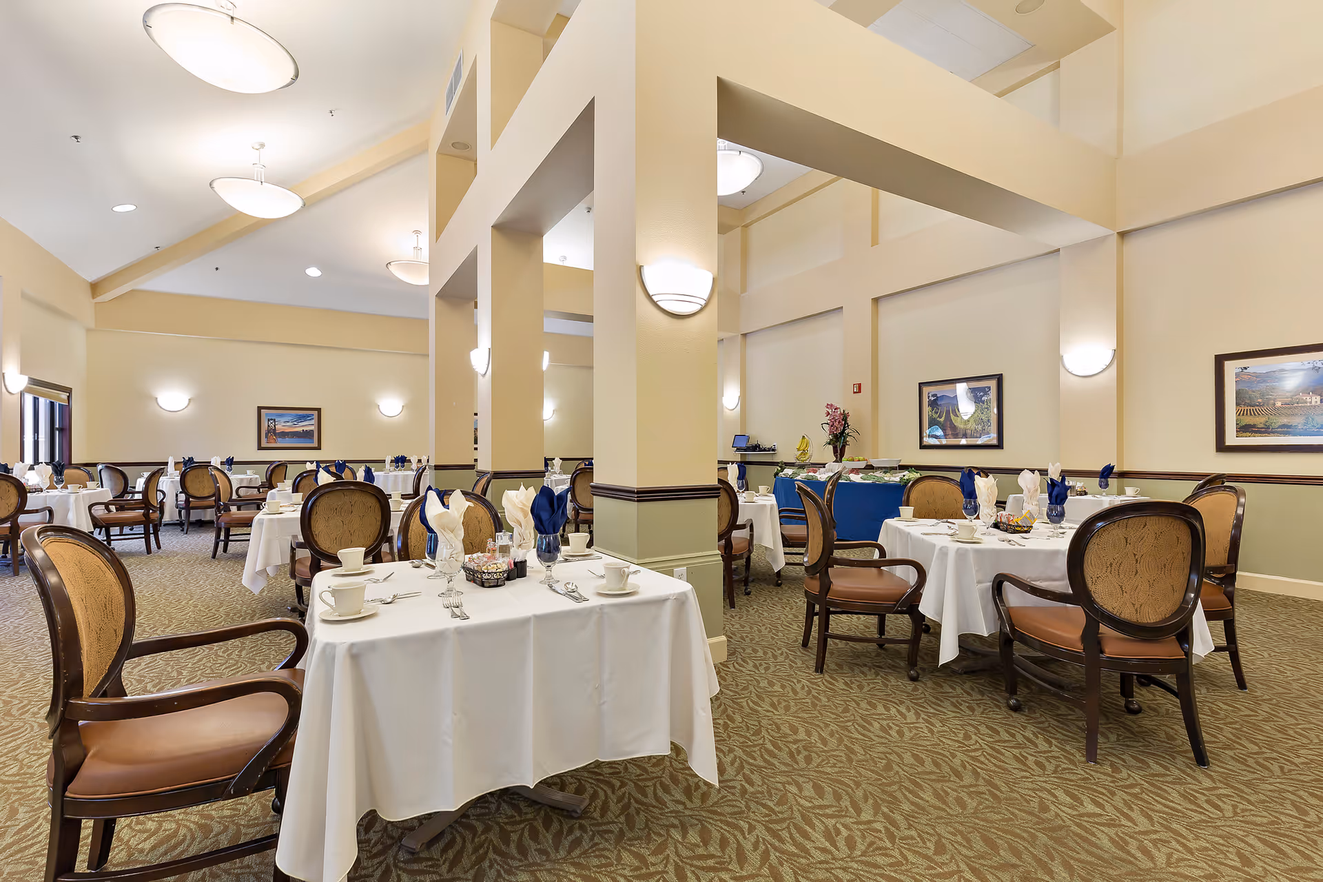 A spacious dining room with multiple round and rectangular tables covered with white tablecloths, set with cups, saucers, silverware, and folded napkins in blue and white. The room has beige walls, carpeted floors with a leaf pattern, and several framed pictures on the walls. There are cushioned wooden chairs around the tables and modern ceiling lights providing illumination.