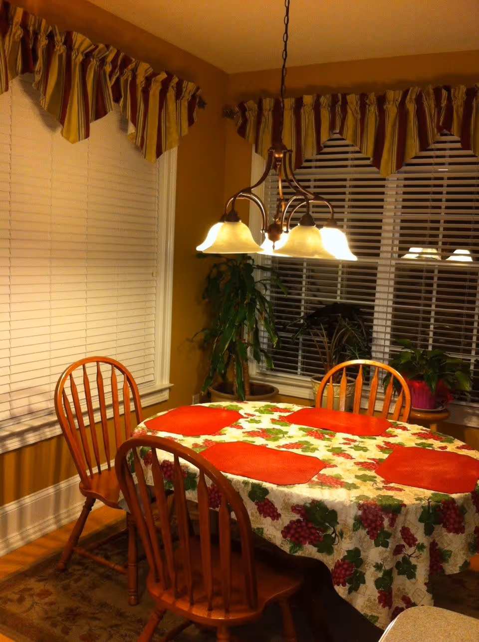 A dining area with a wooden table covered by a tablecloth featuring a grapevine pattern and four red placemats. Four wooden chairs surround the table. Above the table hangs a chandelier with four lights. The room has large windows with white blinds and striped valances. There are potted plants near the windows.