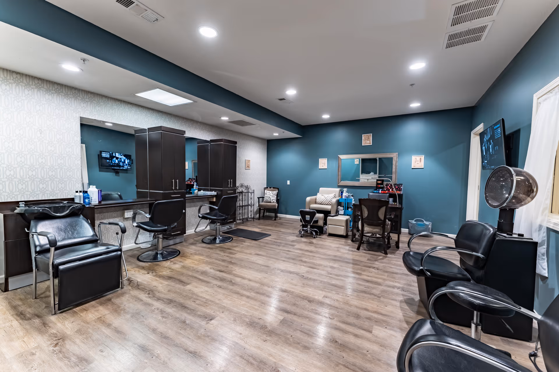 Interior view of a salon area with multiple black salon chairs, mirrors, and cabinets along one wall. The room has wood flooring, teal and patterned wallpaper walls, and ceiling lights. There is a hair dryer station and a small seating area with a beige chair and a table with nail polish bottles.