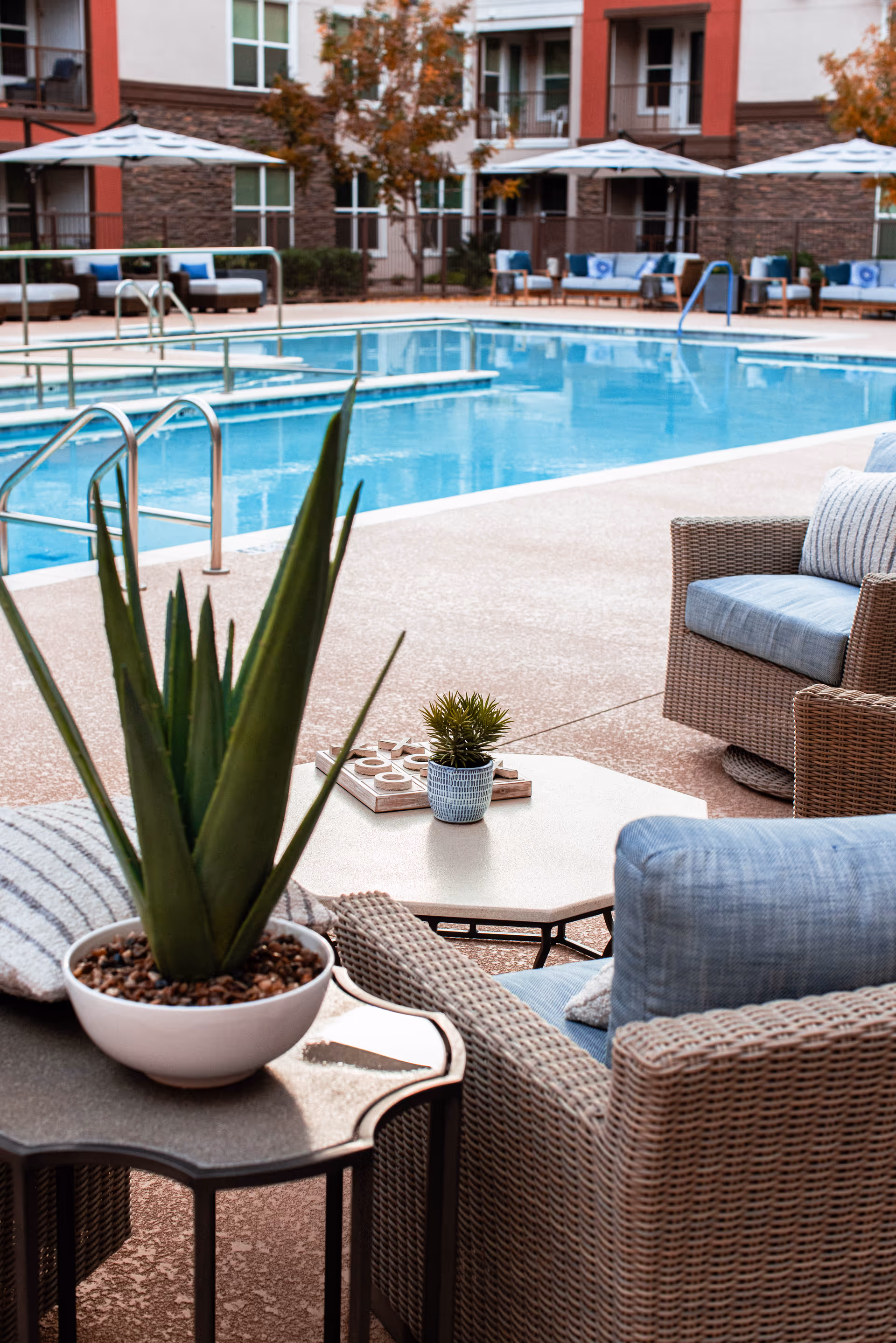 Outdoor pool area with wicker chairs featuring blue cushions, small tables with potted plants, and umbrellas. The pool is surrounded by a patio and a multi-story building with windows and balconies in the background.