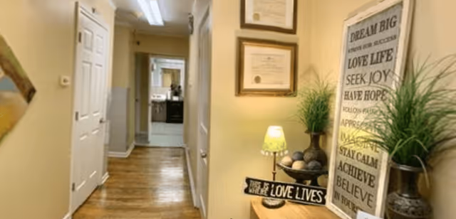 A hallway with wooden flooring and cream-colored walls. On the right side, there is a small table with a lamp, decorative plants, and a sign that reads 'This is where love lives.' Above the table, there are framed certificates and a large framed inspirational quote. The hallway leads to another room with a sink and mirror visible in the background.