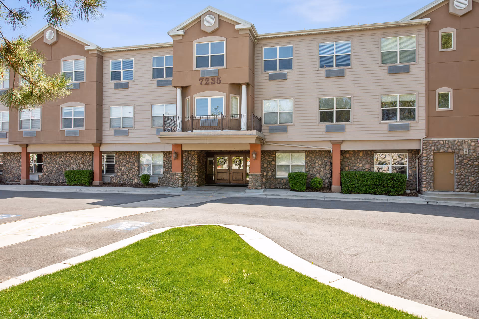 Front exterior view of a three-story senior living facility building with beige and brown siding, stone accents on the lower level, multiple windows, a balcony above the main entrance, and a green lawn in the foreground.