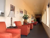 A long indoor hallway lined with red upholstered chairs, side tables with lamps and plants, and windows along one side.