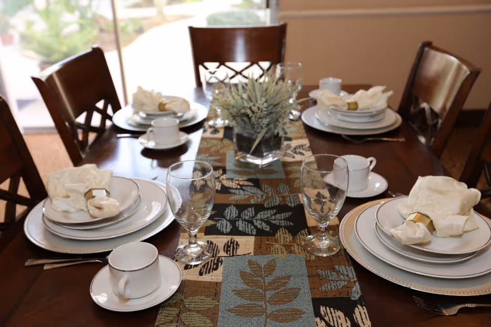 A dining table set for four with white plates, cups, and folded napkins with gold napkin rings. There are two water glasses at each place setting and a decorative table runner with a leaf pattern in the center. A small potted plant is placed in the middle of the table. The room has wooden chairs and a window in the background.