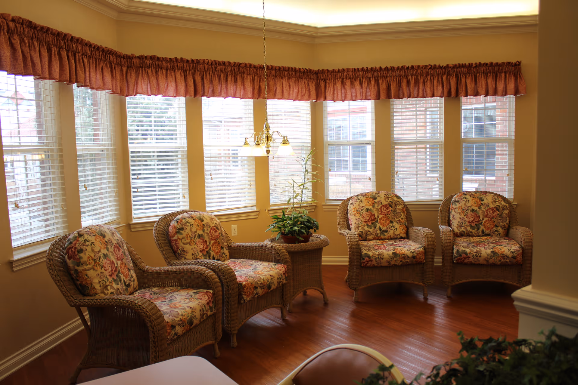 A cozy sitting area with four wicker armchairs featuring floral cushions arranged around a small wicker side table with a potted plant. The room has large windows with white blinds and a red valance, wooden flooring, and a ceiling light fixture hanging above the table.
