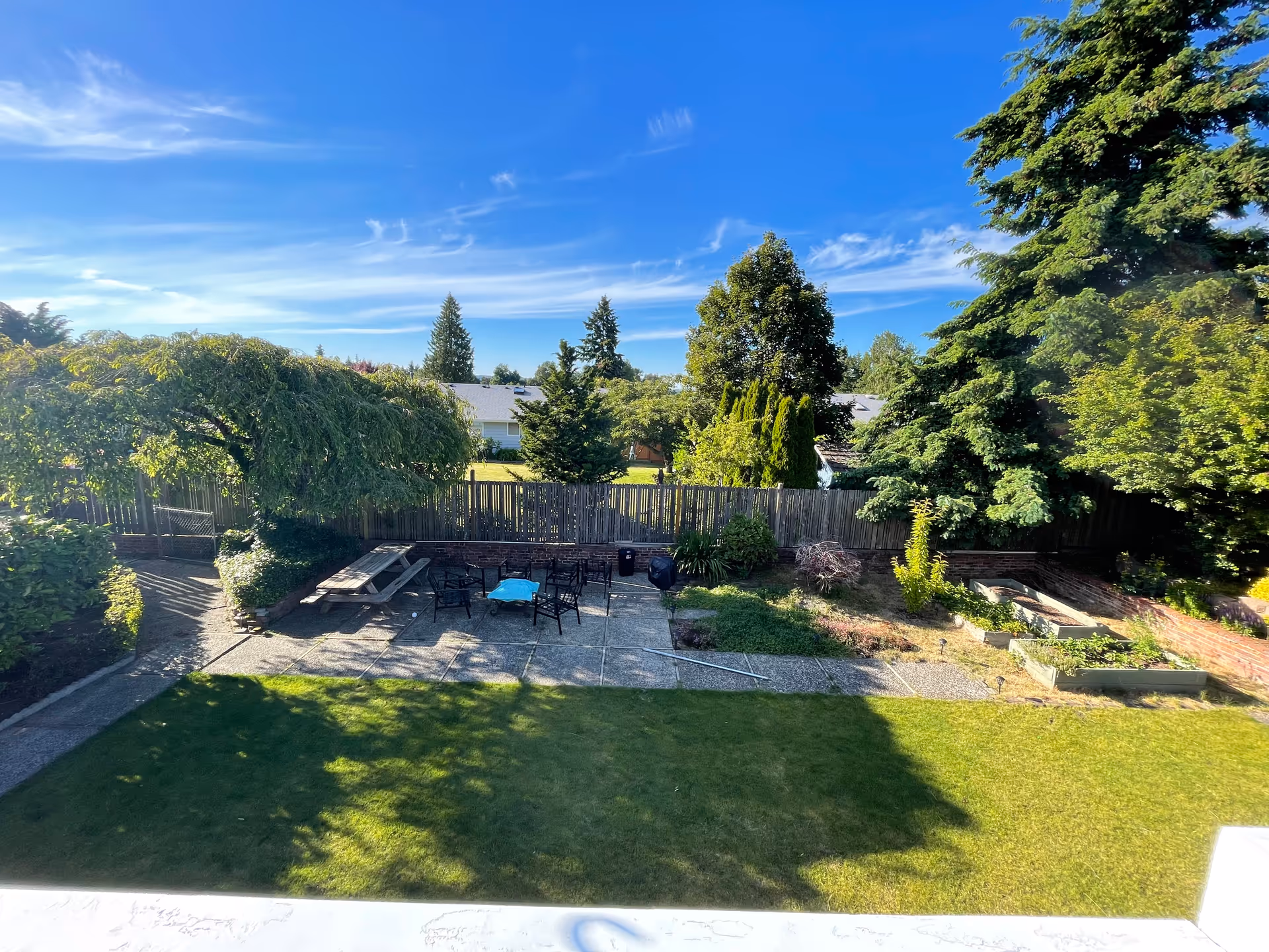 A sunny outdoor garden area with a paved patio featuring a picnic table, several chairs, and a small table with a blue cloth. The garden is surrounded by a wooden fence and lush green trees and shrubs under a clear blue sky.