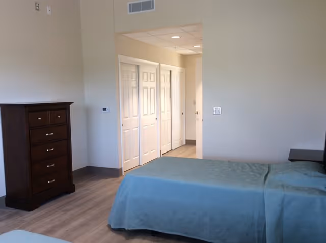 Simple senior living bedroom with twin beds covered in teal bedspreads, a wooden dresser, and a doorway with white closet doors.