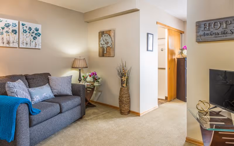 A cozy living room area with a gray couch adorned with several pillows and a blue throw blanket. A side table with a lamp and a floral arrangement sits next to the couch. The walls are decorated with framed artwork and a wooden sign that reads 'HOPE Anchors the Soul Hebrews 6:19'. A flat-screen TV is partially visible on a glass stand to the right. The room has beige carpeting and neutral-colored walls.