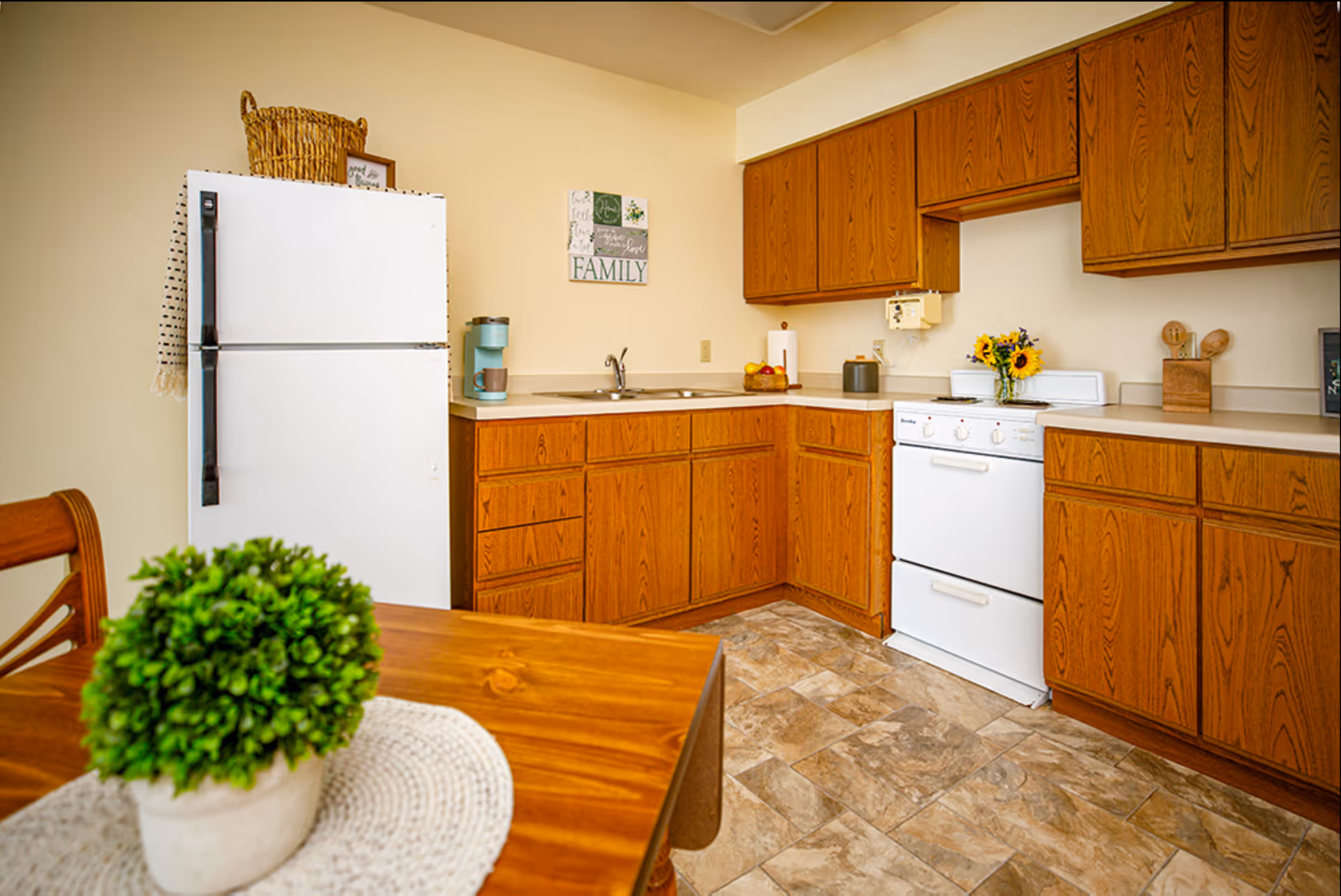 A kitchen with wooden cabinets, a white refrigerator, a white stove, a double sink, and a countertop with a coffee maker, fruit bowl, and a vase of sunflowers. A wooden dining table with a green potted plant on a woven placemat is visible in the foreground.