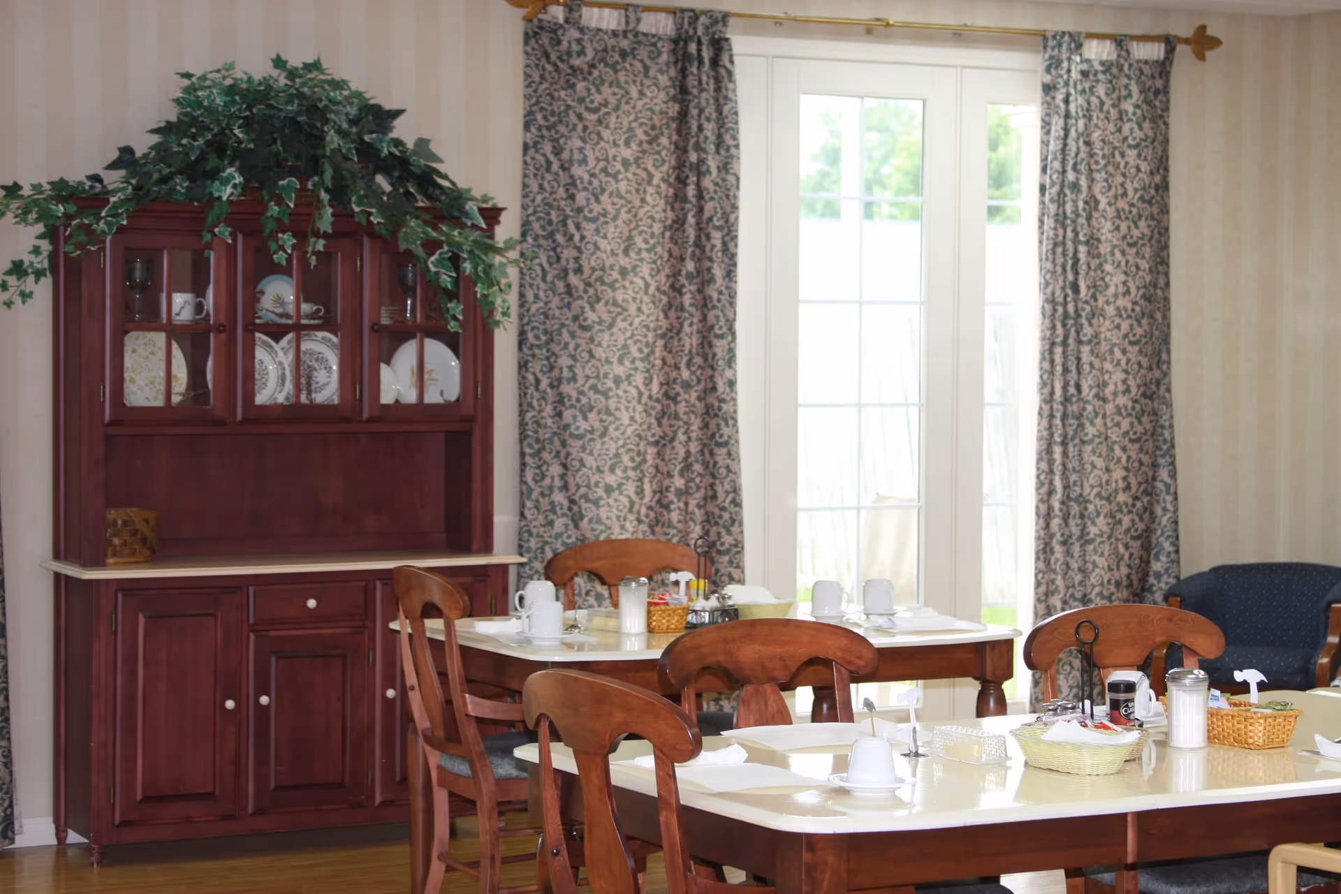 A dining room with wooden tables and chairs set for a meal, including cups, napkins, and condiments. There is a wooden cabinet with glass doors displaying plates and cups, topped with a green leafy plant. Behind the tables are large windows with patterned curtains, and part of a blue upholstered chair is visible on the right.