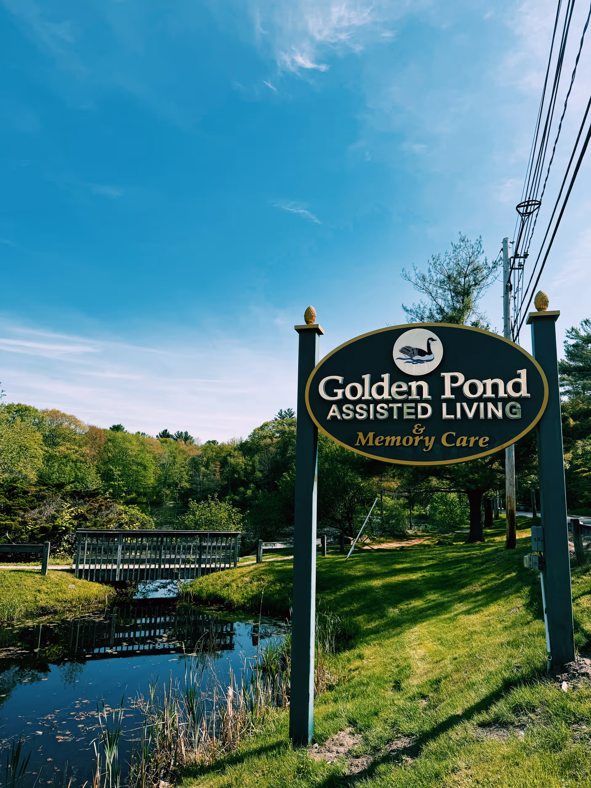 A sign for Golden Pond Assisted Living & Memory Care stands on a grassy area next to a small pond with a wooden bridge. Trees and a clear blue sky are visible in the background.