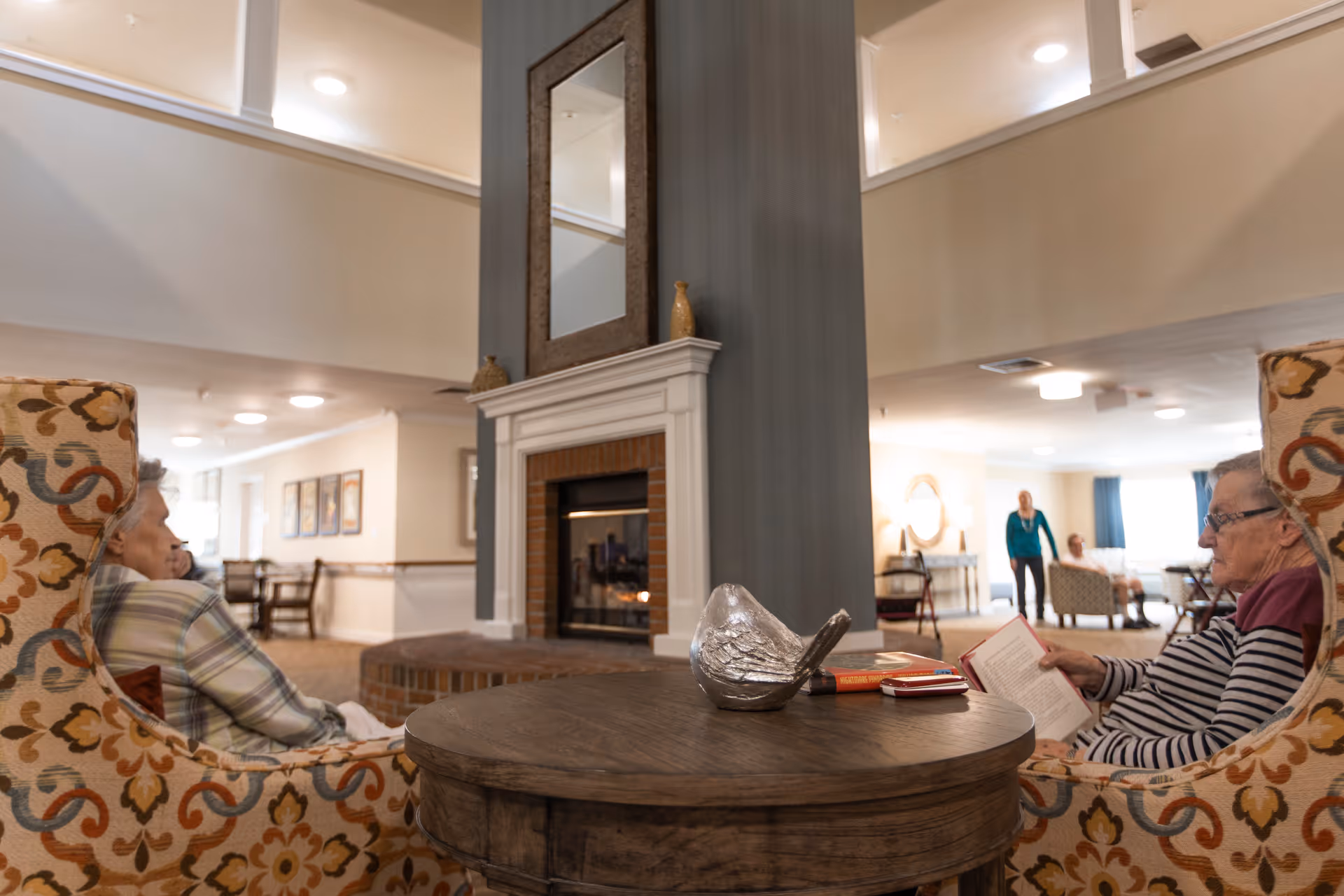 Two elderly women sitting in patterned armchairs facing each other near a fireplace with a mirror above it in a spacious, well-lit senior living common area. One woman is reading a book while the other looks ahead. In the background, other residents and a caregiver are visible in the lounge area.