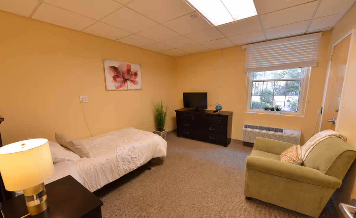 A cozy bedroom in Birchwood Suites with a single bed covered in a white textured bedspread, a bedside table with a lamp, a green armchair with a pillow and blanket, a black dresser with a small TV and decorative items, a window with white blinds, and a painting of a red flower on the beige wall.