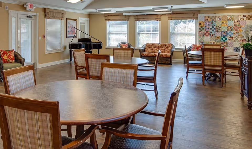 A bright communal lounge with round dining tables and chairs, sofas, a piano, and large windows.