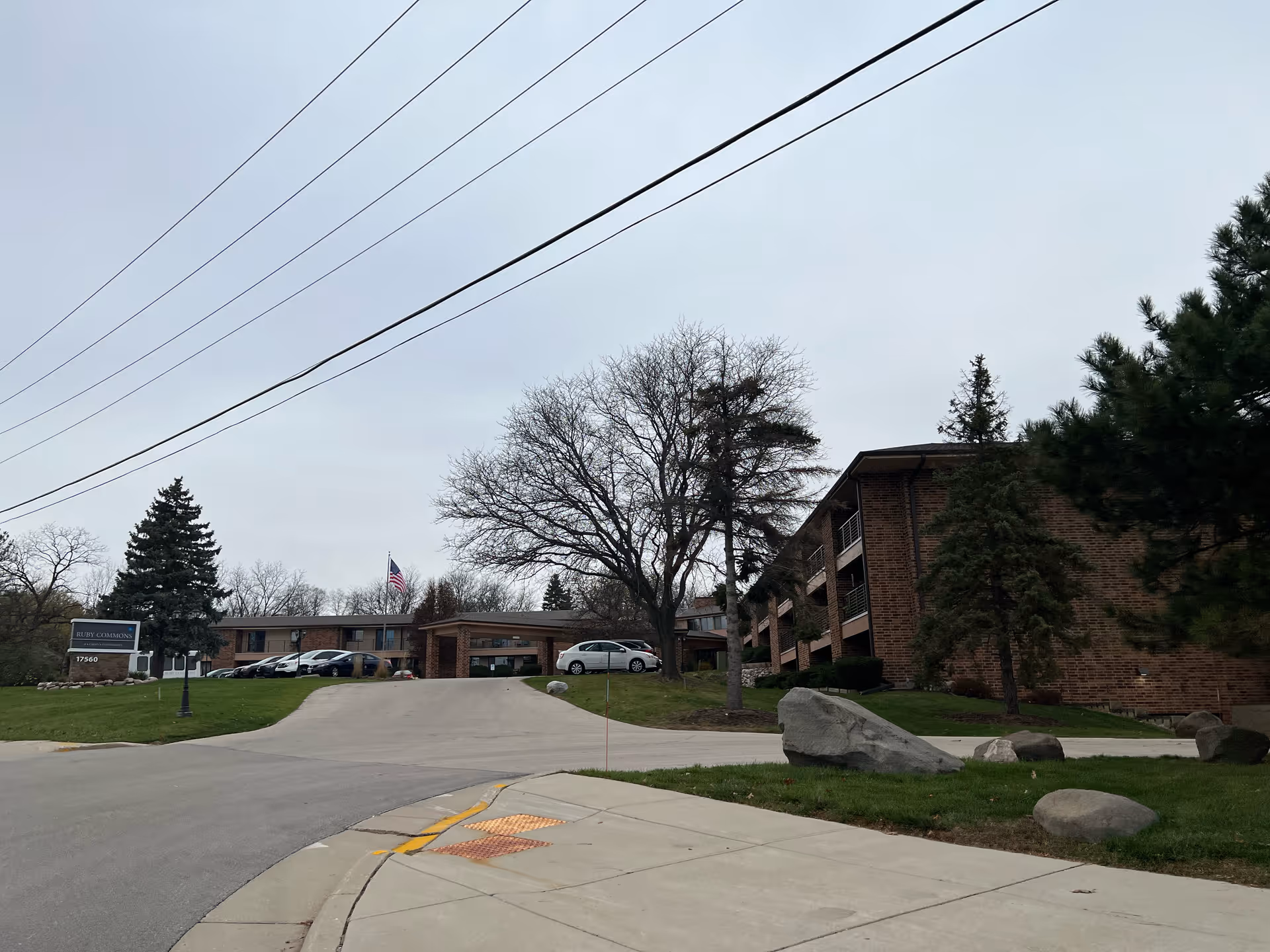 Exterior view of Ruby Commons senior living facility showing a brick building with balconies, a driveway leading to the entrance, several parked cars, trees without leaves, and an American flag on a flagpole.