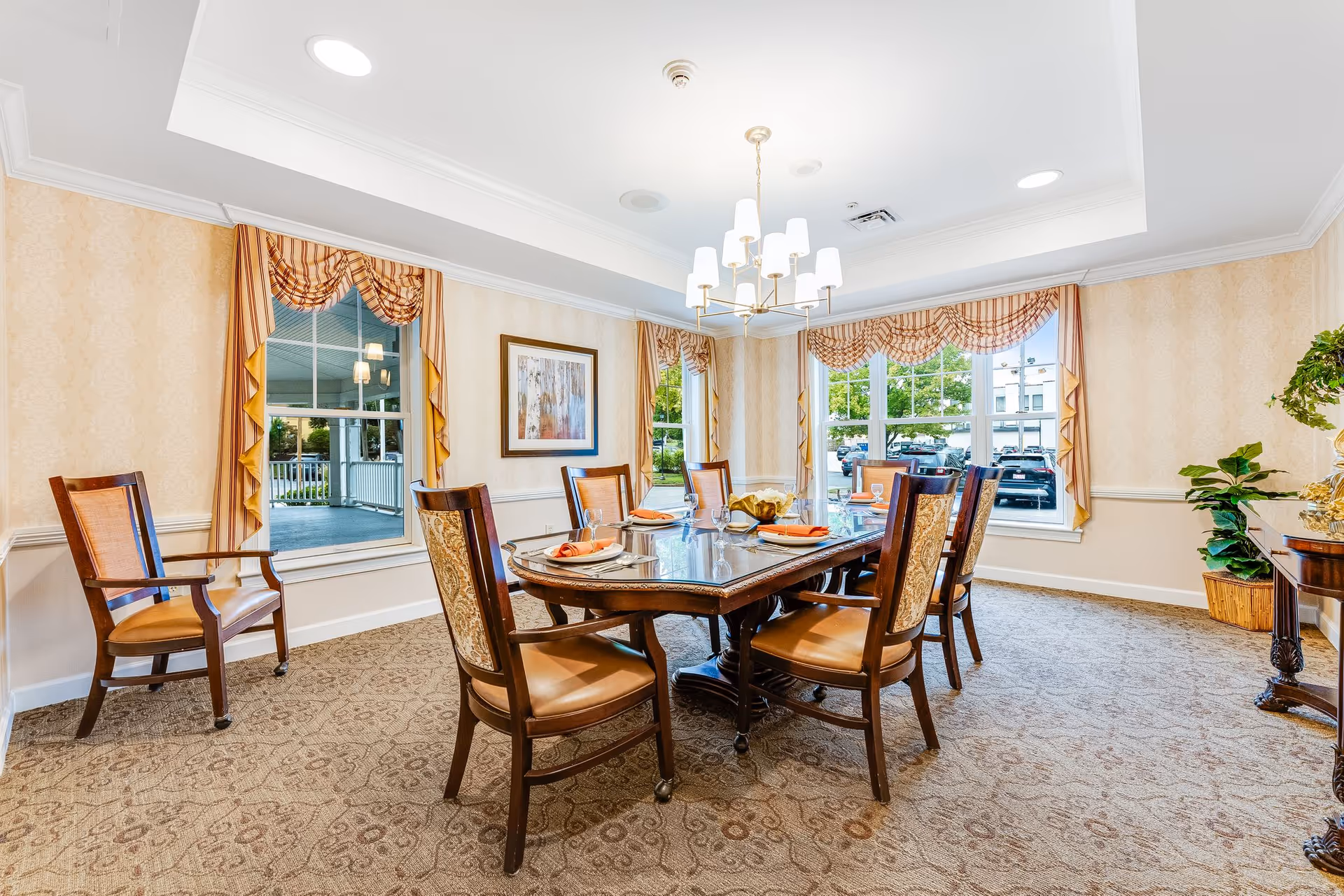 A well-lit dining room with a large rectangular wooden table set for six people. The table has glassware, plates, and orange napkins. The room features patterned carpet, beige wallpaper with a subtle design, and three large windows with striped and ruffled curtains. A chandelier with multiple lights hangs above the table, and there is a framed artwork on the wall. A wooden side table with a plant and decorative items is visible on the right side.