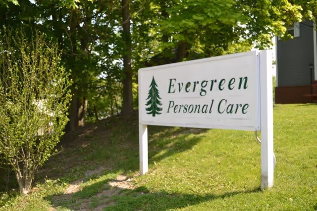 A white yard sign that reads "Evergreen Personal Care" with a pine tree logo on a grassy lawn near trees and a building.