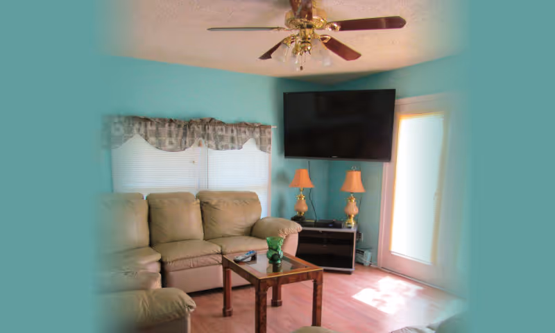 A cozy living room with a beige leather sectional sofa, a wooden coffee table with a green decorative item and a remote control on it, two table lamps with beige shades on a small stand, a wall-mounted flat-screen TV, and a ceiling fan with lights. The walls are painted light blue, and there is a window with blinds and a valance, as well as a glass door letting in natural light.