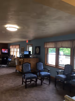 Interior view of a senior living facility common area with several upholstered chairs arranged near large windows. A television is on in the background with a person sitting nearby. The room has carpeted flooring, light blue walls, and a ceiling light fixture.