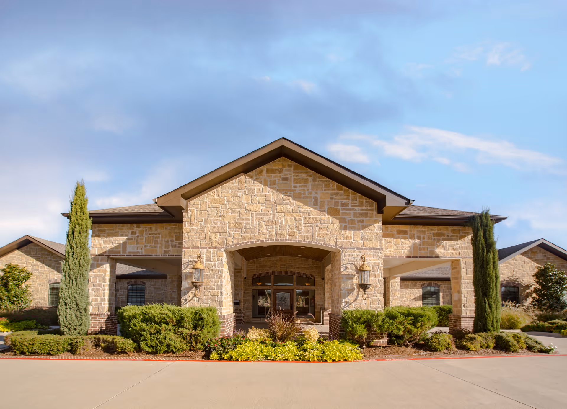 Front exterior view of Sandy Lake Rehabilitation and Care Center, a single-story building with stone facade, large entrance with an archway, flanked by tall narrow trees and well-maintained shrubs and plants under a partly cloudy sky.