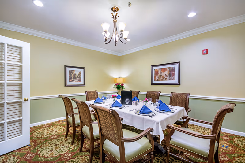A dining room with a rectangular table set for eight people. The table is covered with a white tablecloth and has blue folded napkins, glassware, and silverware. The room has patterned carpet, light green and beige walls with two framed paintings, a lamp on a side table, and a chandelier hanging from the ceiling. A white door with glass panels is partially open on the left side.