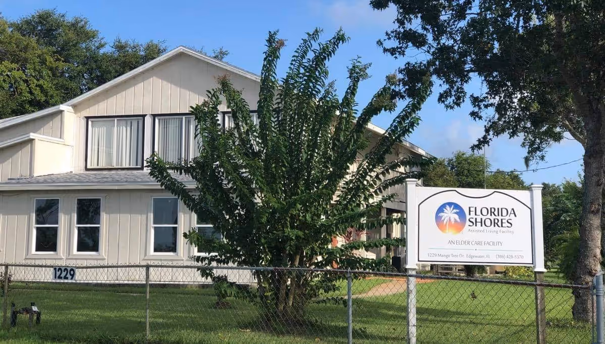 Front view of a light‑colored two-story building with a lawn, chain‑link fence, and a 'Florida Shores' assisted living sign.