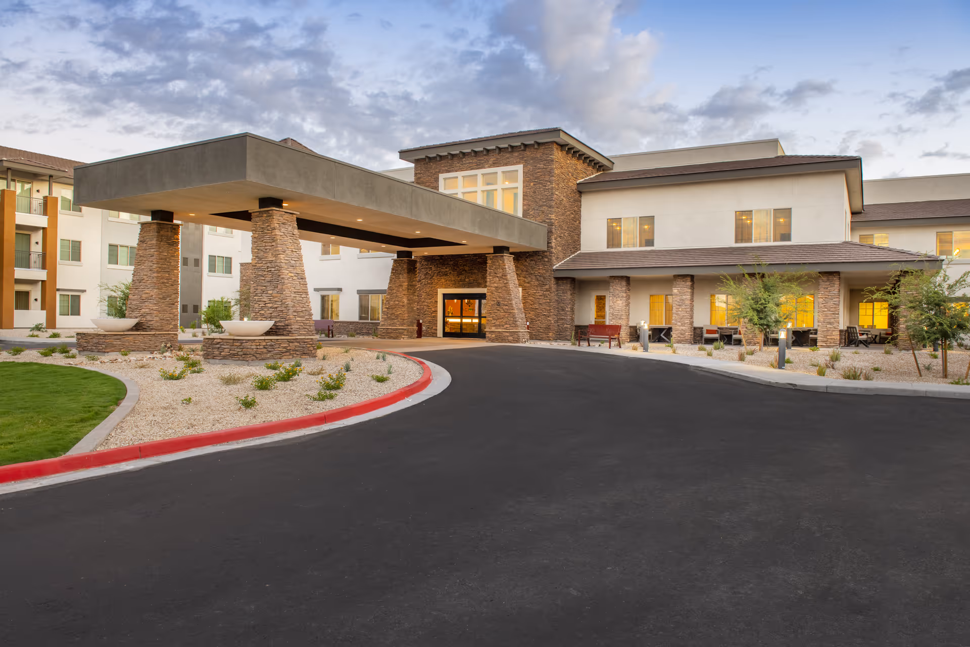 Exterior view of a modern senior living facility building with stone pillars supporting a covered entrance. The building has multiple windows, some with lights on inside, and landscaped areas with small plants and trees. The sky is partly cloudy during dusk.