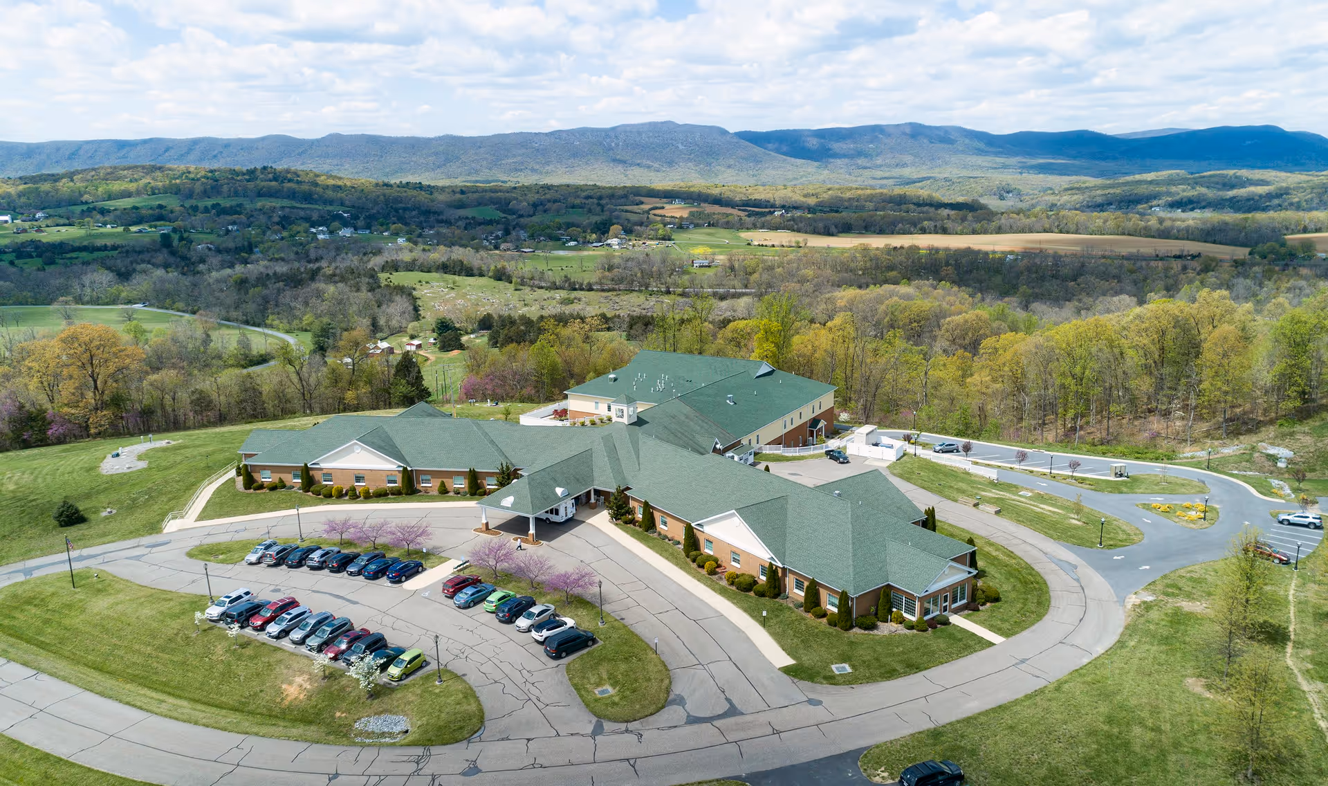 Aerial view of Hidden Springs Senior Living facility surrounded by green lawns and trees, with a parking lot filled with cars in front. The building has green roofs and is situated in a rural area with hills and forests in the background.