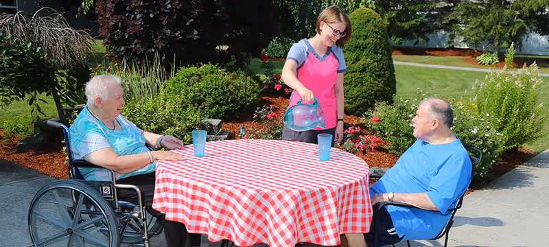 An elderly woman in a wheelchair and an elderly man sitting at a round table with a red and white checkered tablecloth outdoors. A caregiver wearing a pink apron is pouring water into a glass on the table. The setting is a garden area with green bushes, flowers, and a paved walkway.
