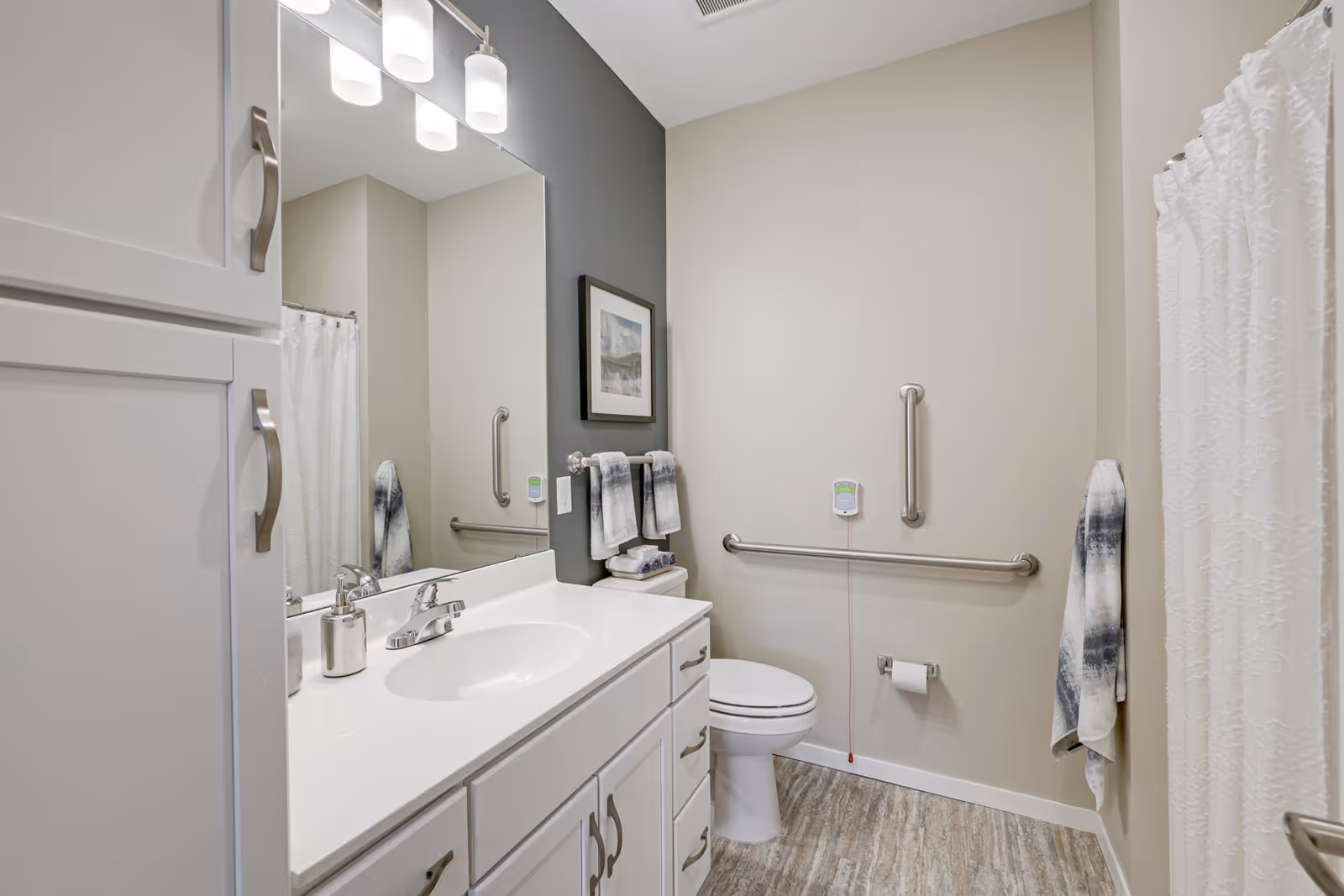 A clean and modern bathroom with a white vanity and sink, a large mirror above the sink, a toilet with grab bars on the wall for accessibility, a towel rack with folded towels, and a shower with a white curtain. The walls are painted in neutral tones and the floor has a wood-like finish.