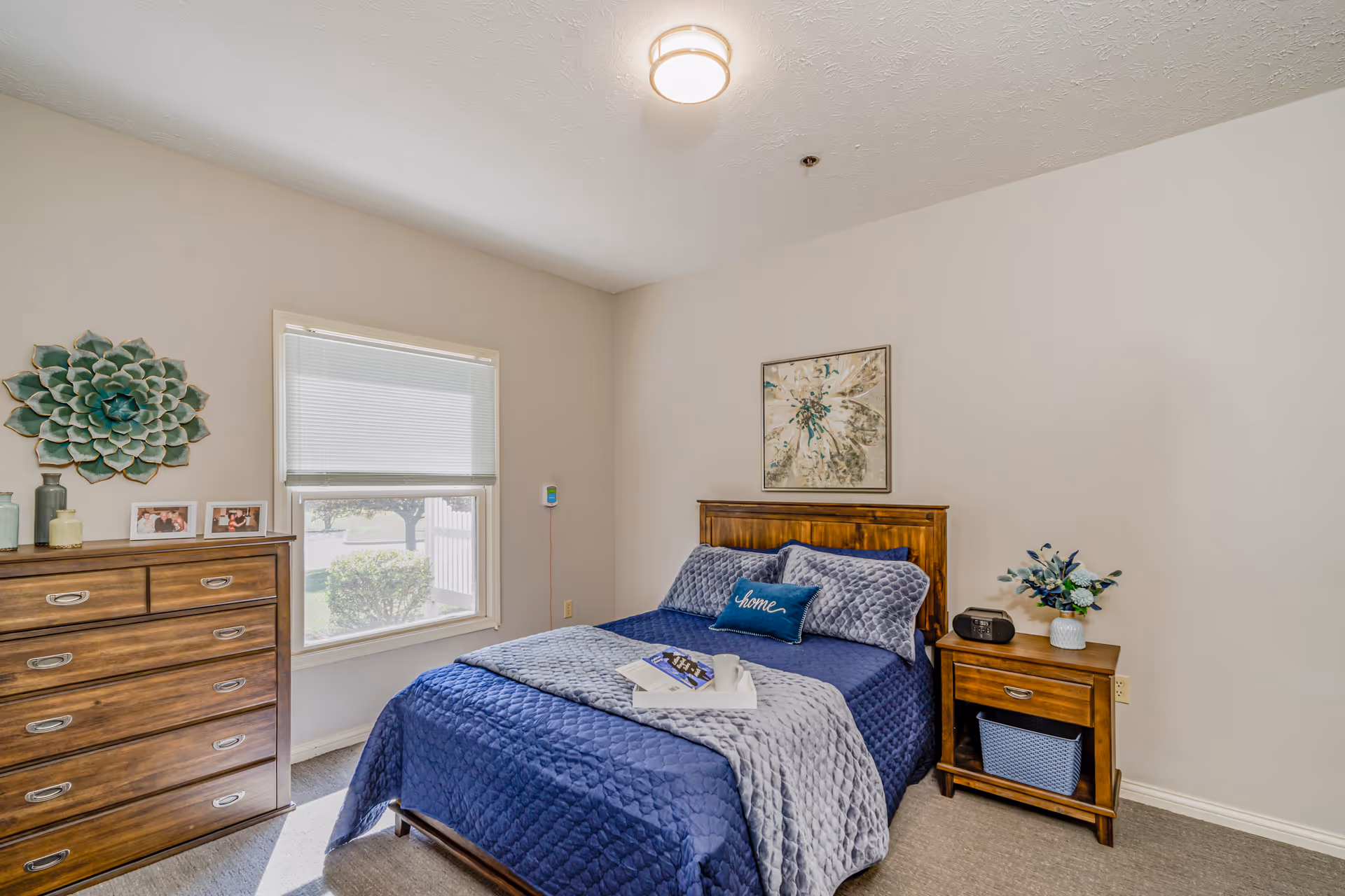 A cozy bedroom with a wooden bed frame and blue bedding, including a quilted blanket and pillows. A tray with a book and a cup is placed on the bed. Next to the bed is a wooden nightstand with a small clock and a vase with flowers. On the left side of the room is a wooden dresser with decorative vases and framed photos. A window with blinds lets in natural light, and a large decorative flower wall art hangs above the dresser. The walls are painted light beige, and the carpet is light gray.