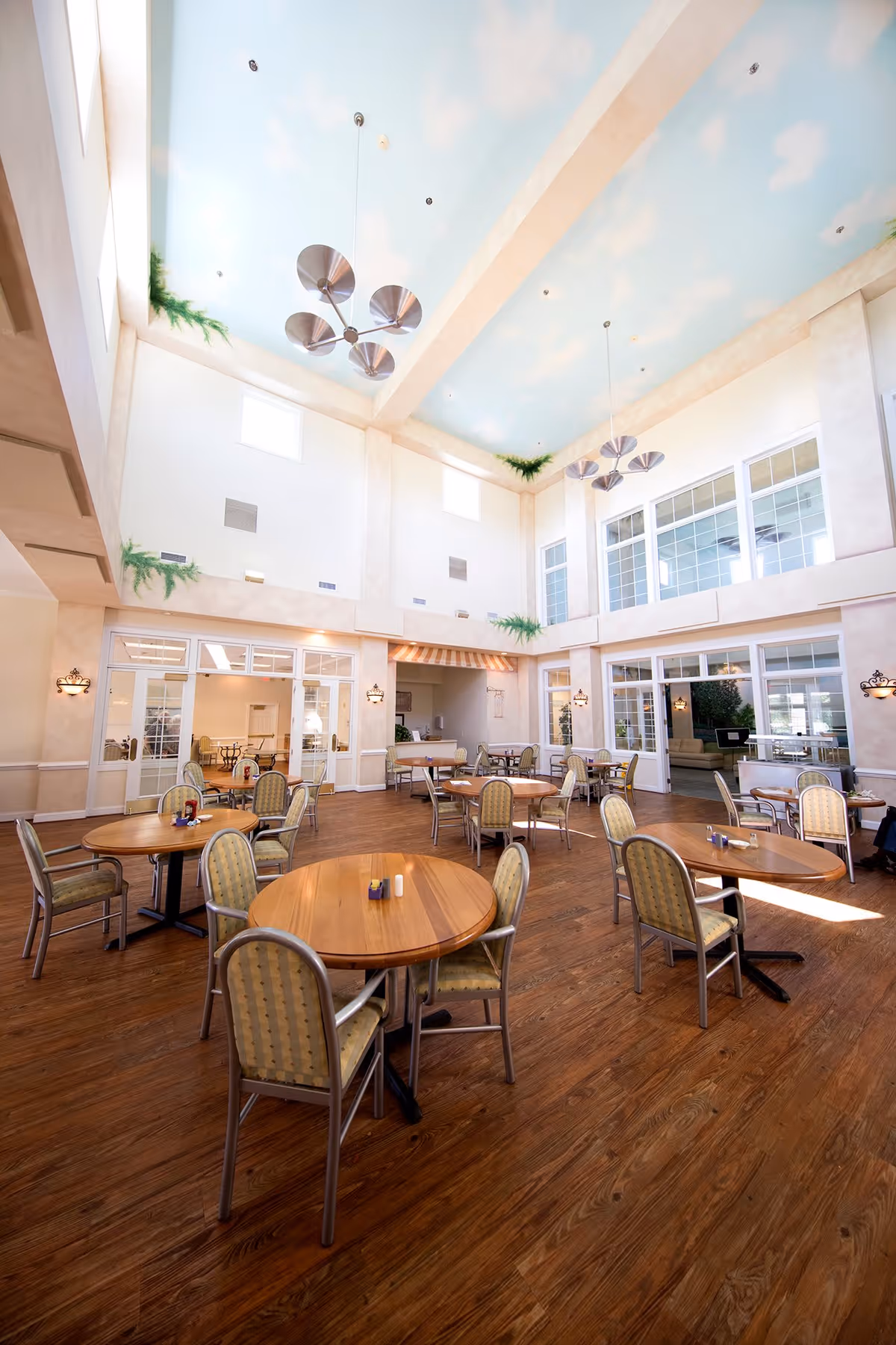 A spacious dining room with multiple round wooden tables and cushioned chairs arranged neatly on a wooden floor. The room features high ceilings painted with a sky and clouds motif, large windows allowing natural light, and modern hanging light fixtures. There are plants placed on ledges near the ceiling and wall sconces providing additional lighting.