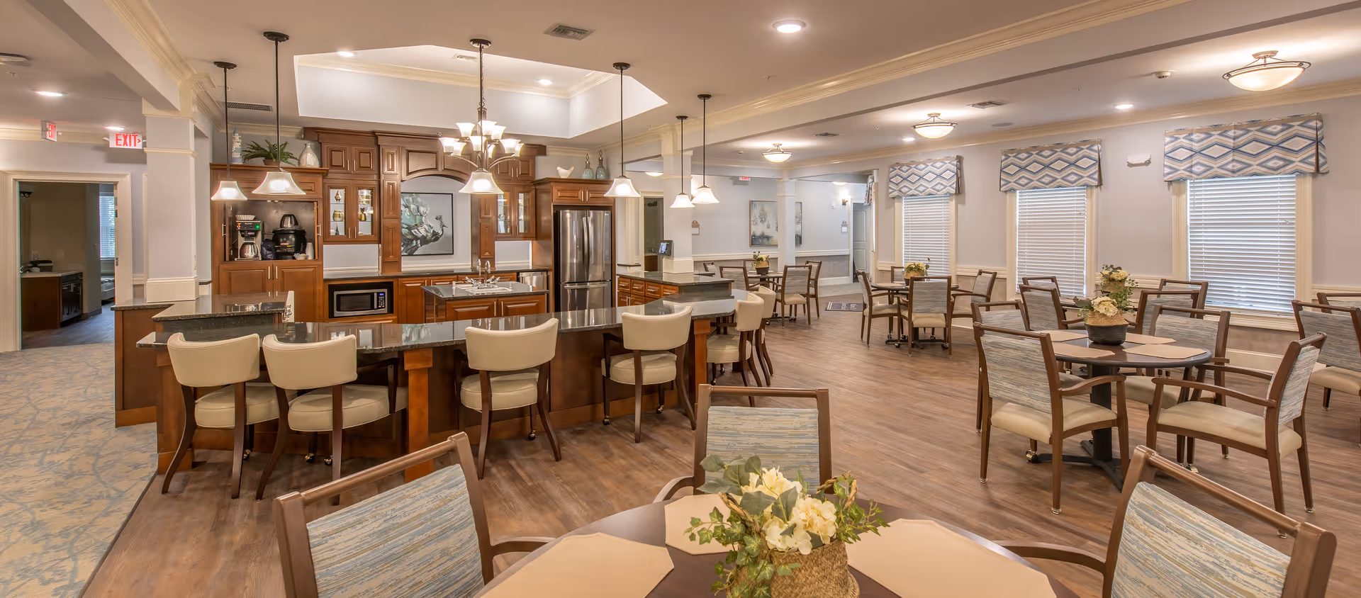 A spacious dining area with multiple round tables and chairs, each table decorated with a floral centerpiece. The room features large windows with patterned valances, wood flooring, and soft lighting. In the background, there is a kitchen area with dark wood cabinetry, a granite countertop bar with cream-colored bar stools, and stainless steel appliances.