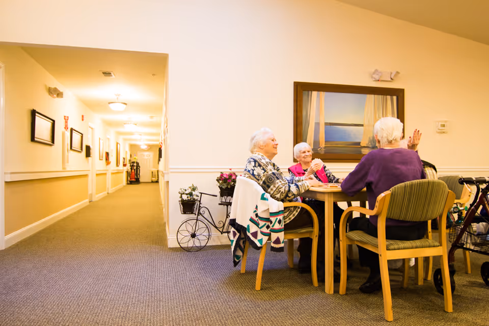 A group of elderly people sitting around a wooden table in a well-lit hallway or common area of a senior living facility, engaged in conversation or an activity. The walls are light-colored with framed pictures, and there is a decorative plant stand with flowers nearby. A walker is visible next to one of the chairs.