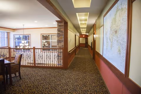Interior view of a senior living facility hallway with patterned carpet and a large map on the right wall. To the left, there is a railing overlooking a dining area with tables and chairs, illuminated by hanging light fixtures.