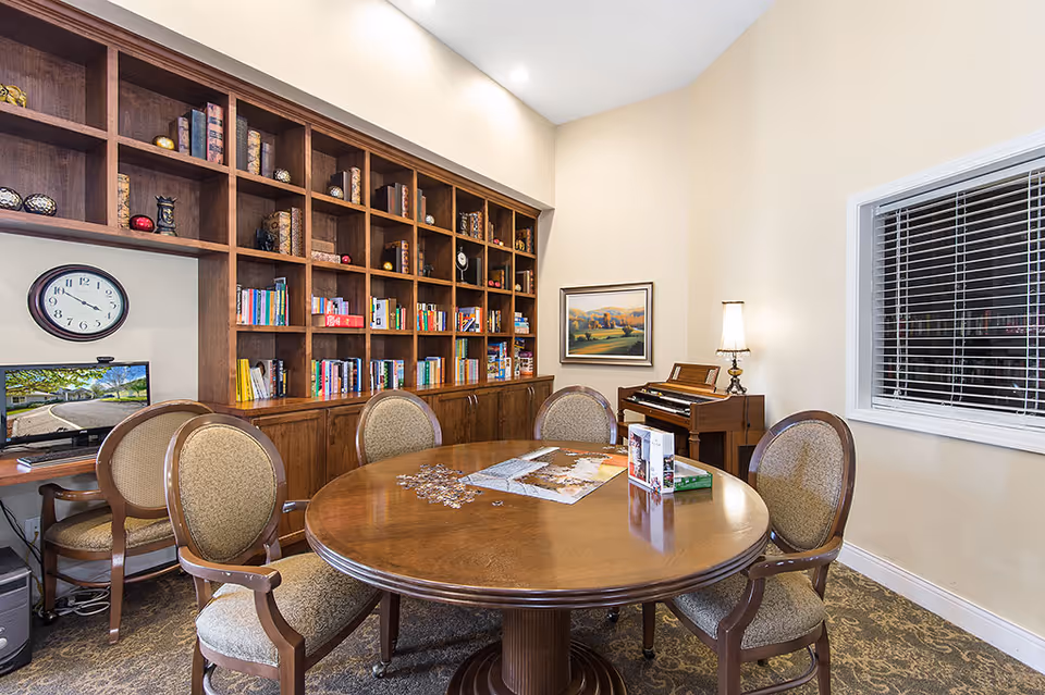 A cozy room with a round wooden table surrounded by five upholstered chairs. On the table, there is a partially completed jigsaw puzzle and some puzzle boxes. Behind the table is a large wooden bookshelf filled with books and decorative items. A wall clock and a computer monitor are visible on the left side. On the right side, there is a small piano with a lamp on top and a framed landscape painting on the wall. A window with blinds is on the right wall.