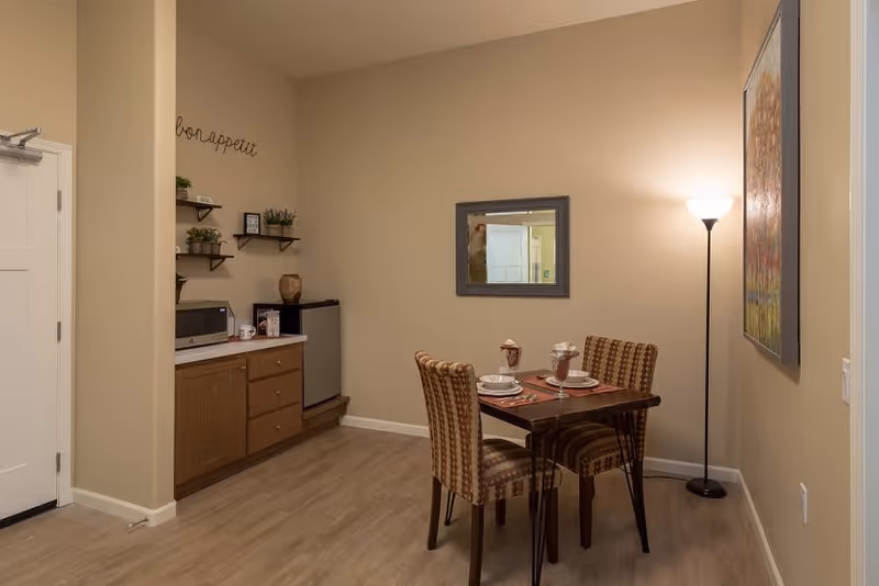 A small dining area in a senior living facility with a wooden table set for two, two patterned chairs, a floor lamp, a wall mirror, and a colorful painting. To the left, there is a kitchenette area with wooden cabinets, a microwave, a small refrigerator, and wall shelves with decorative plants and items. The walls are painted beige and the floor has light wood laminate.