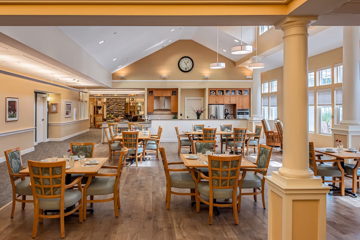 A spacious dining room with multiple wooden tables and chairs arranged neatly. The room features high ceilings with hanging light fixtures, large windows allowing natural light, and a kitchen area with stainless steel appliances in the background. The walls are painted in warm beige tones, and there are decorative columns and framed pictures on the walls.