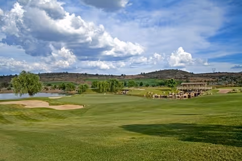 A scenic view of a golf course with well-maintained green grass, sand bunkers, a small pond, and a wooden bridge. The background features rolling hills under a partly cloudy blue sky.