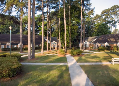 Courtyard with paved walkways, benches and tall pine trees surrounded by single-story brick residential buildings.