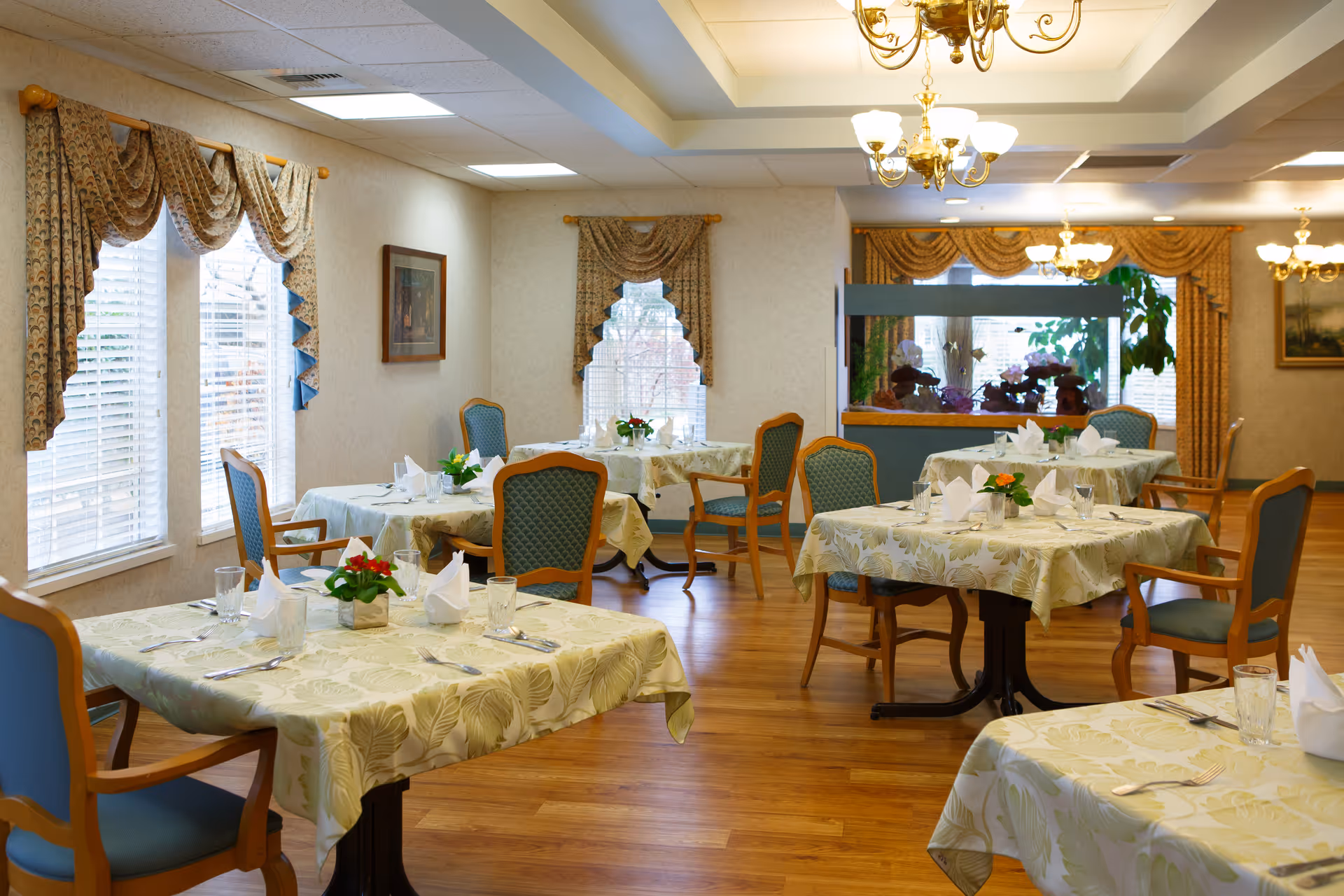 Bright dining room with several tables set with napkins, flowers, and upholstered chairs.