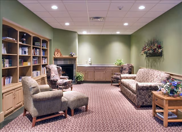 A cozy living room area with patterned carpet, two upholstered armchairs, a matching sofa, a wooden coffee table with a flower arrangement, built-in wooden bookshelves filled with books, a fireplace, and green walls decorated with plants and flowers.