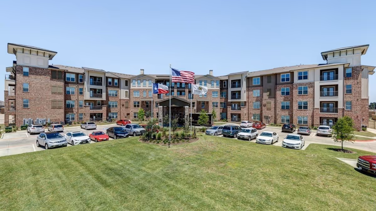Exterior view of a four-story senior living facility building with brick and beige siding, multiple windows and balconies, a well-maintained green lawn in front, and several parked cars. Three flagpoles with the American flag, Texas state flag, and a facility flag are centered near the entrance.