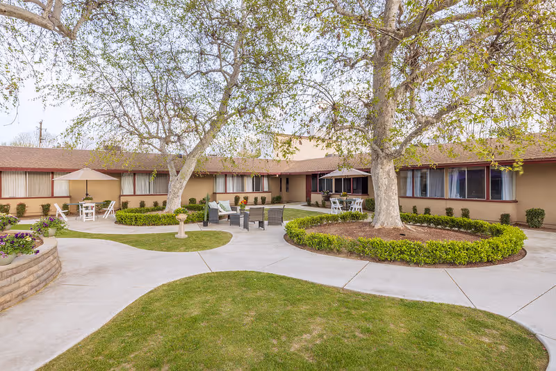 Outdoor courtyard area of a nursing and rehabilitation center featuring a circular concrete walkway, green grass, two large trees with trimmed bushes around their bases, and several seating areas with chairs and tables under umbrellas.