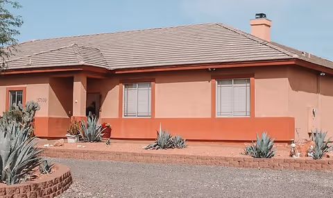 Single-story residential building with a tiled roof and peach-colored exterior walls, surrounded by desert landscaping including agave plants and gravel. The building has multiple windows with blinds and a chimney on the right side.