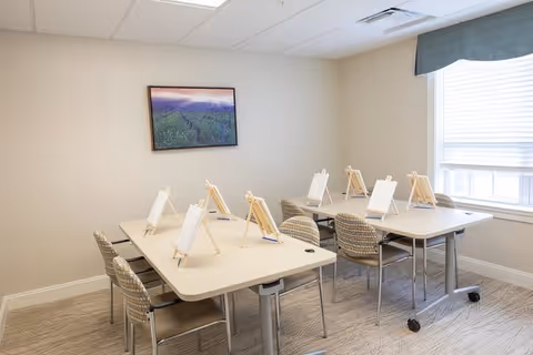 A bright room with two rectangular tables arranged in a U-shape, each table holding small easels with blank canvases. There are eight patterned chairs around the tables. A large window with blinds lets in natural light, and a framed landscape photograph hangs on the beige wall.