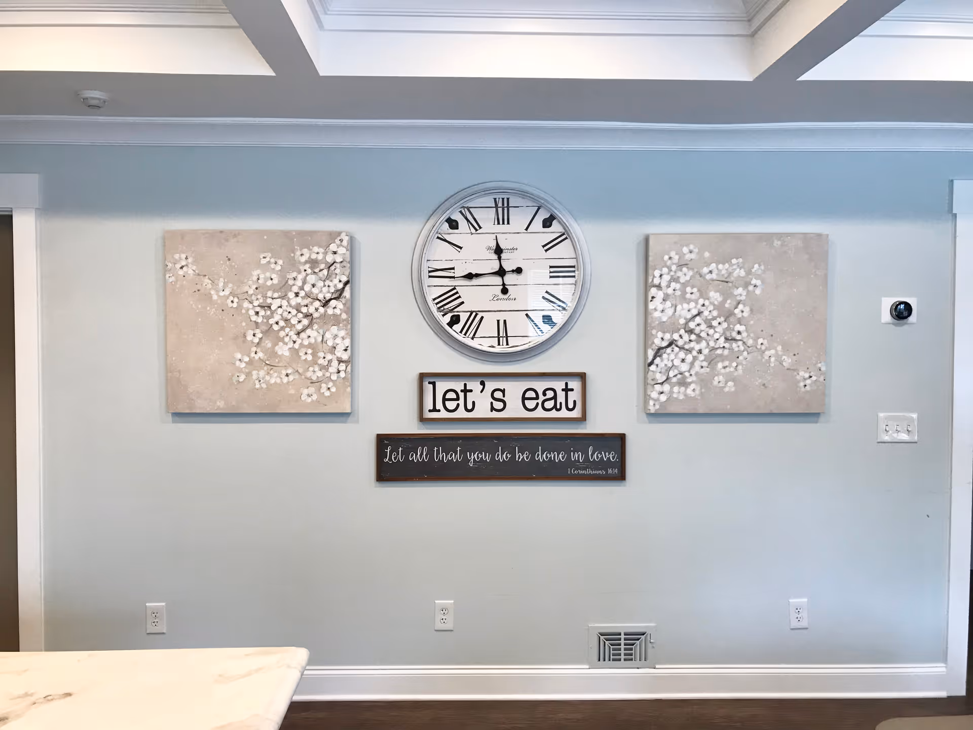 Interior wall with a large round clock showing 8:45, two square floral paintings on either side, and two wooden signs below the clock. One sign says 'let's eat' and the other says 'Let all that you do be done in love. 1 Corinthians 16:14'. The wall is light blue with white trim and a marble countertop is partially visible at the bottom left.