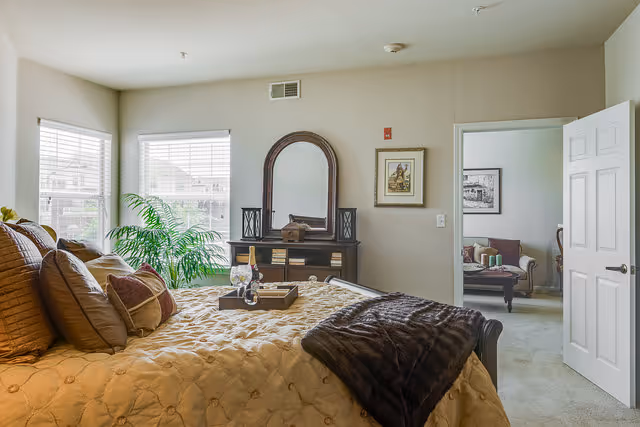 A cozy bedroom with a large bed covered in a beige quilt and multiple pillows. A dark wooden dresser with a large arched mirror sits against the wall, flanked by two decorative lanterns. A tray with a bottle of wine and two glasses is placed on the bed. Two windows with white blinds allow natural light to fill the room. A green potted plant is near the windows. An open door reveals a glimpse of a living room with a sofa and framed artwork on the walls.